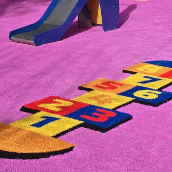 Zona infantil con tobogán azul sobre suelo de césped artificial color rosa, decorado con un diseño de rayuela en colores vivos; ideal para ilustrar ejemplos de instalar césped artificial en zonas infantiles.