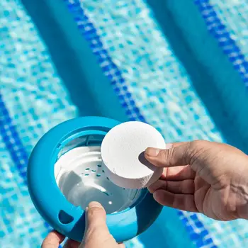 Primer plano de una mano introduciendo una pastilla de cloro en un dispensador flotante sobre una piscina, junto a una zona pavimentada lista para la instalación de césped artificial Turfgrass.