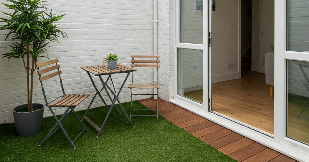 Imagen en la que se ve un patio interior con césped artificial, mesa y sillas de madera, una planta en maceta y con unas puertas correderas de cristal.