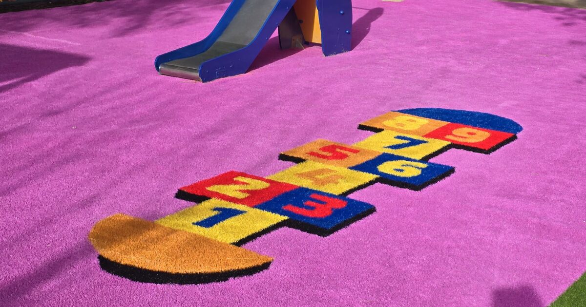 Zona infantil con tobogán azul sobre suelo de césped artificial color rosa, decorado con un diseño de rayuela en colores vivos; ideal para ilustrar ejemplos de instalar césped artificial en zonas infantiles.
