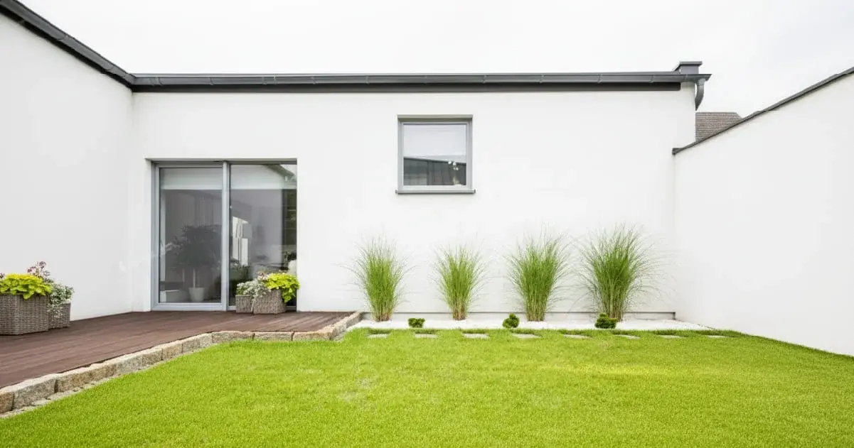 Modern backyard with white house, wooden deck, sliding glass door, green lawn, and ornamental grasses in white gravel.