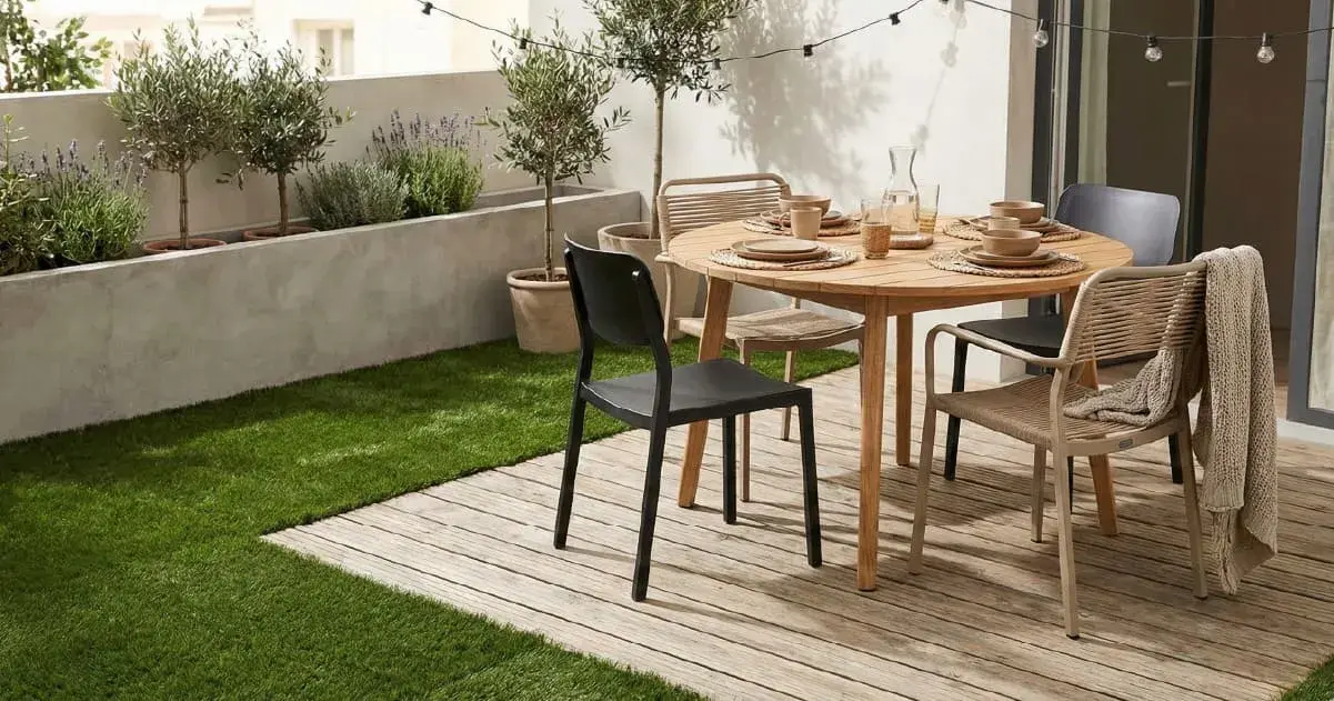 Modern outdoor dining area featuring a round wooden table set for four with mixed black and beige woven chairs on a wooden deck. The space is bordered by artificial grass and a concrete planter with olive trees and lavender under overhead string lights.