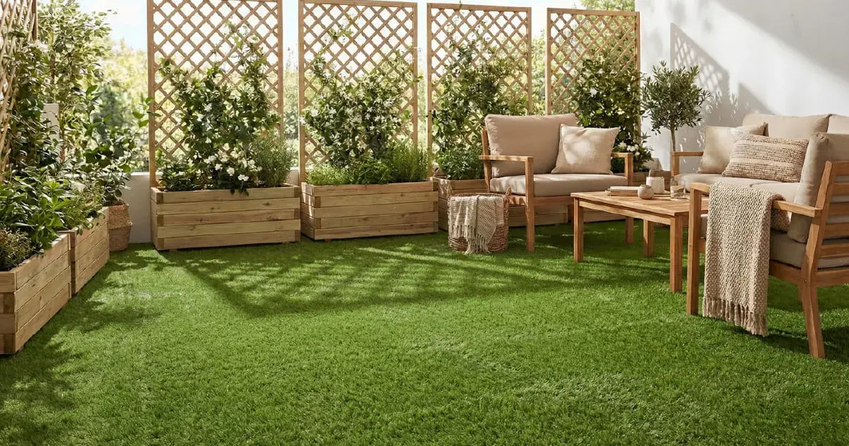 A sun-drenched outdoor terrace featuring artificial grass, wooden lounge furniture with neutral cushions, and large wooden planters with trellises holding lush green plants and white flowers.