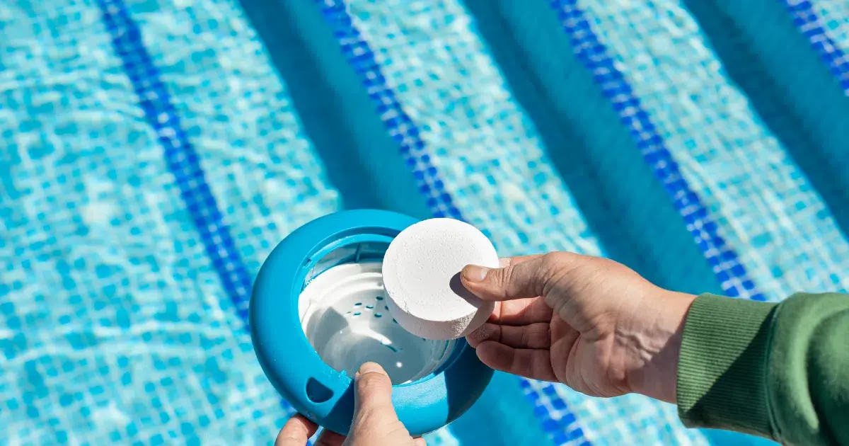 Primer plano de una mano introduciendo una pastilla de cloro en un dispensador flotante sobre una piscina, junto a una zona pavimentada lista para la instalación de césped artificial Turfgrass.