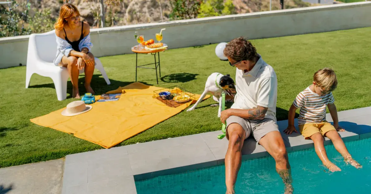 Una familia con su perro relajándose en una zona de ocio exterior con césped artificial Turfgrass junto a una piscina moderna.