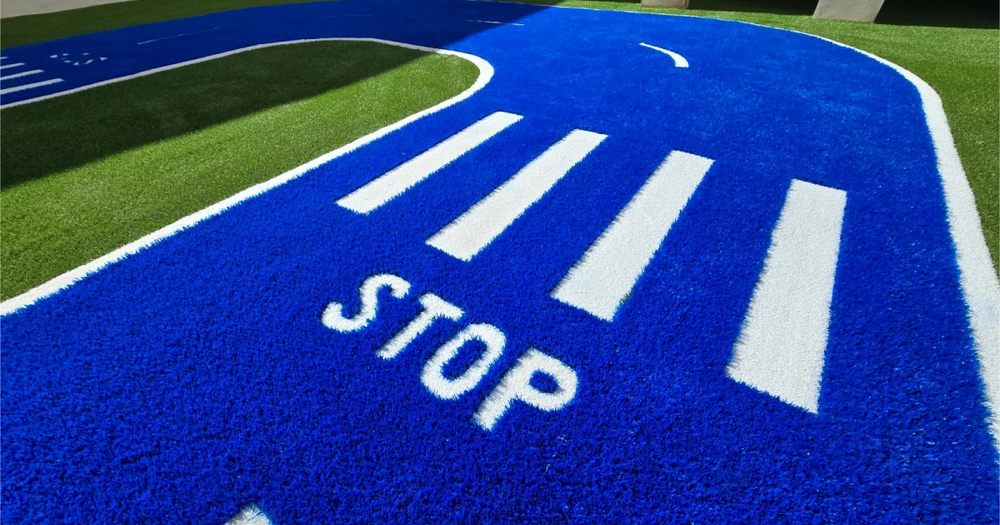 Zona infantil con césped artificial instalado en forma de carretera azul, señales de tráfico y líneas blancas, ejemplo de instalar césped artificial en zonas infantiles.