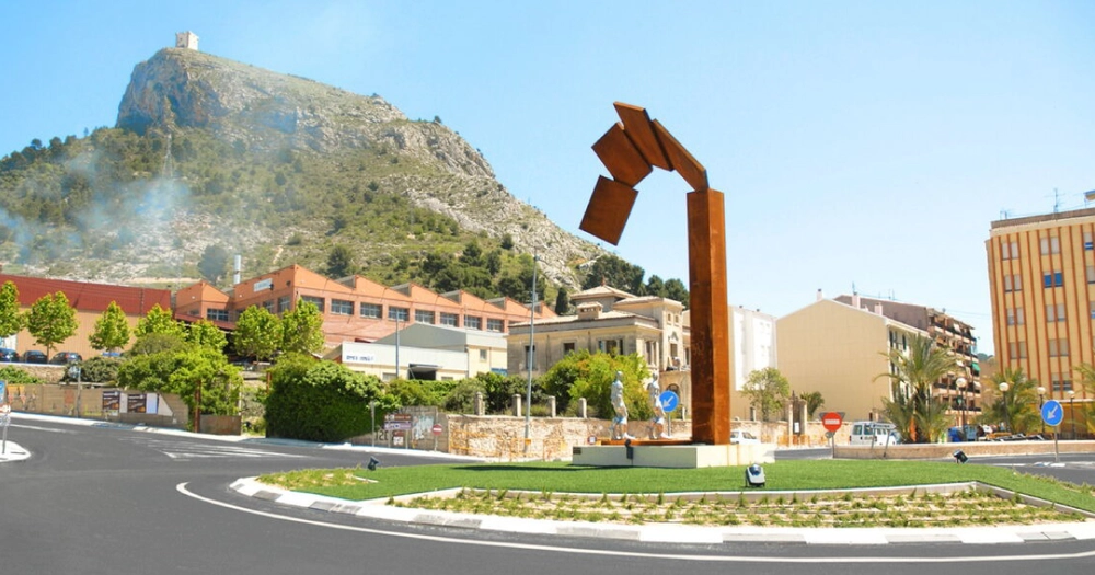 Imagen en la que podemos ver una rotonda de una ciudad con una estructura abstracta y también vemos otro ejemplo de utilizar césped artificial en zonas públicas.